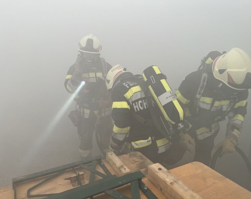 Drei Feuerwehrleute in Schutzausrüstung arbeiten in einem nebligen Bereich, einer hält eine Taschenlampe. Sie bereiten sich auf einen Rettungseinsatz vor.