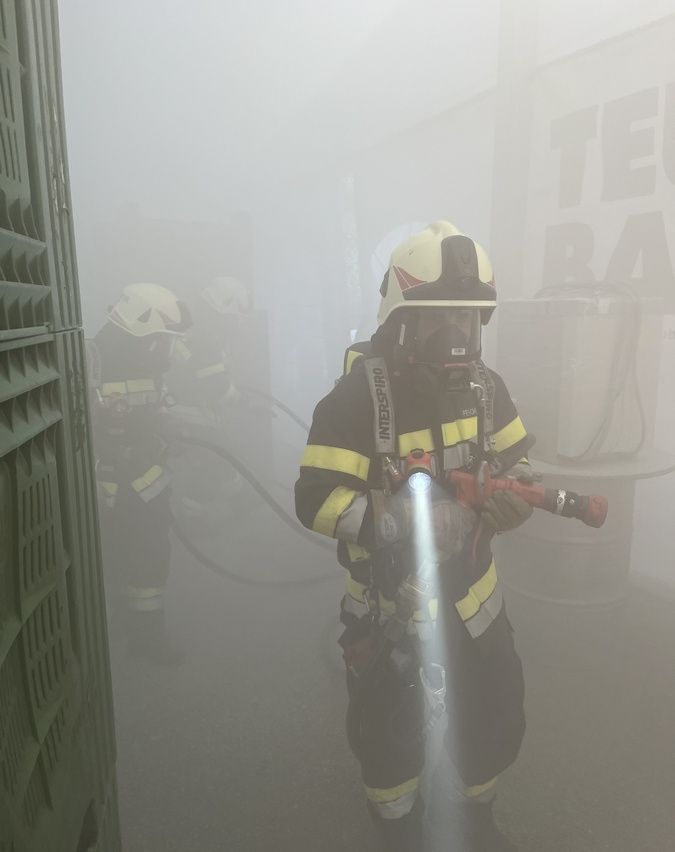 Feuerwehrleute in Schutzausrüstung navigieren durch dichten Nebel. Einer führt mit einer Taschenlampe, während andere folgen. Im Hintergrund sind die Buchstaben 'TE' in großen Lettern zu sehen.