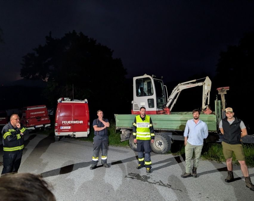 Fünf Männer in Uniformen stehen nachts in einer Gruppe vor Notfallfahrzeugen. Ein großer Bagger ist in der Nähe geparkt.