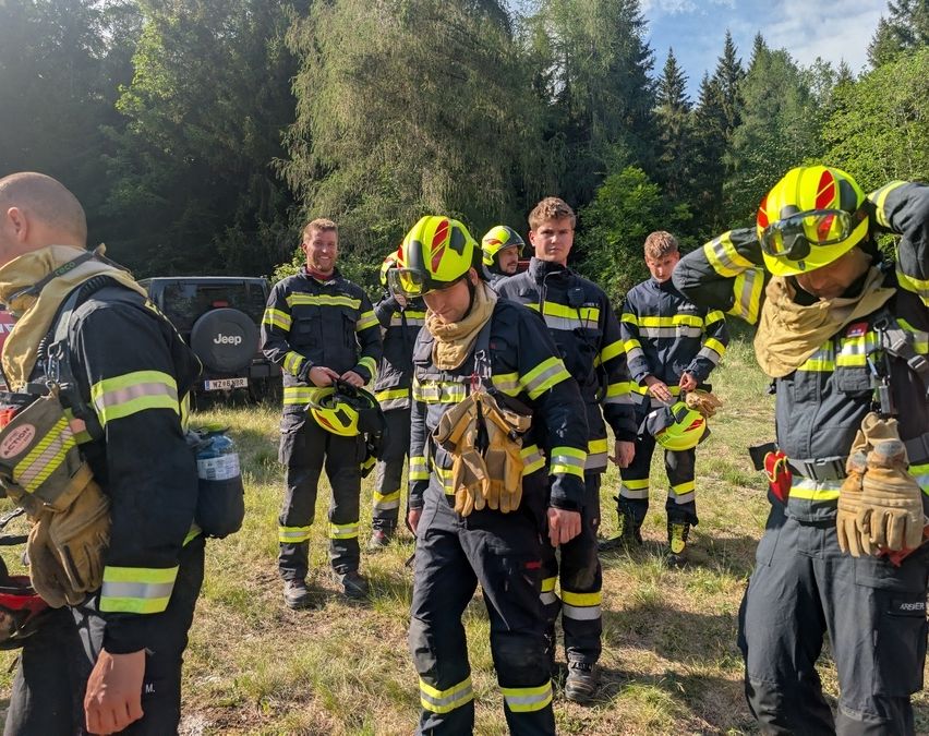 Feuerwehrleute in voller Ausrüstung stehen auf einem Feld, einer justiert einen Helm, ein anderer hält einen Feuerlöscher. Ein Jeep und Bäume im Hintergrund.