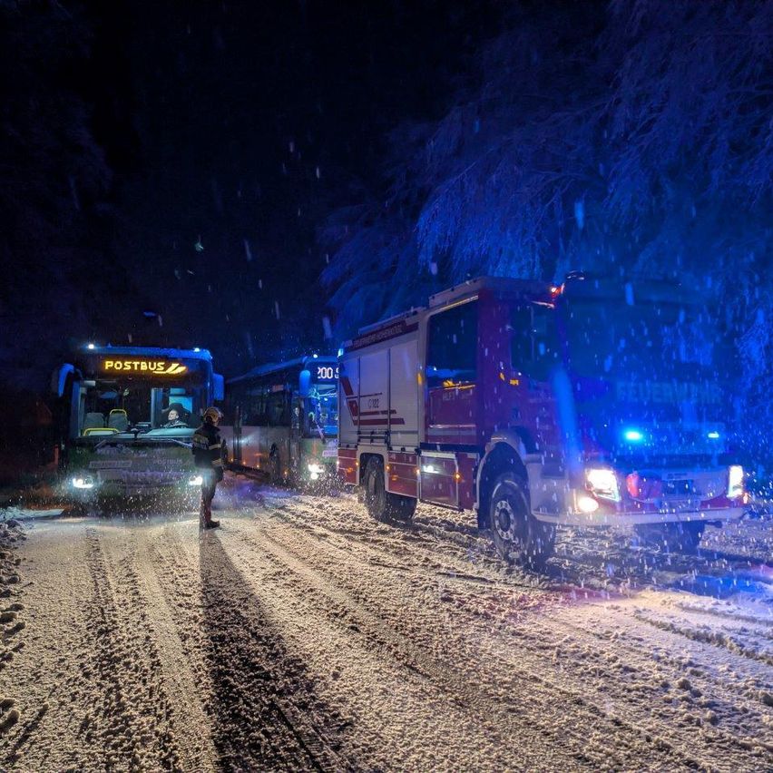 Eine verschneite Straße bei Nacht mit zwei Bussen und einem Feuerwehrwagen, bei einem Bus steht eine Person davor und der Feuerwehrwagen hat seine Lichter eingeschaltet.
