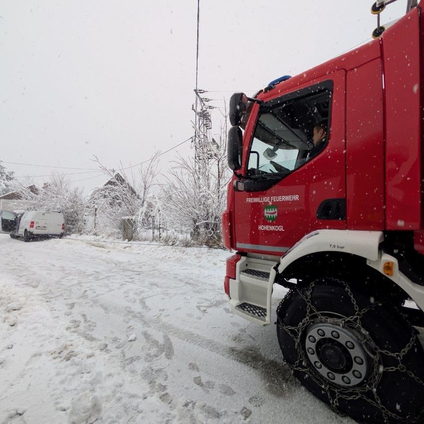 Ein roter Feuerwehrwagen mit Ketten an den Reifen steht im Schnee. Dahinter ist ein weißer Lieferwagen geparkt, und ein Baum ist im Hintergrund.