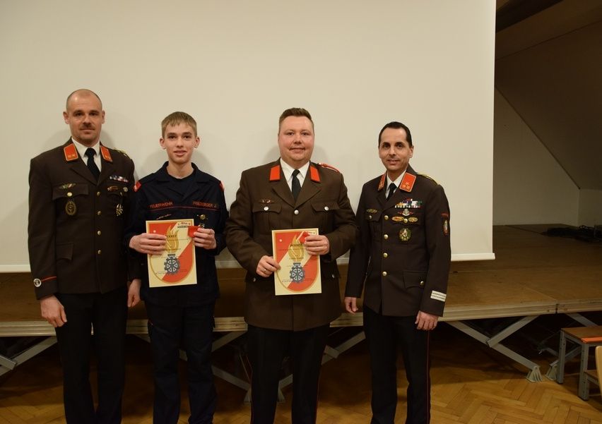 Vier Männer in Militäruniformen, zwei halten Urkunden, stehen in einer formellen Umgebung mit einer weißen Wand und einer hölzernen Bühne im Hintergrund.