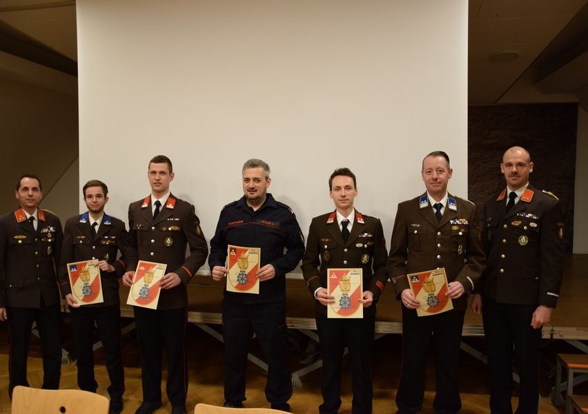 Sieben Männer in Militäruniformen stehen zusammen und halten Auszeichnungen. Hinter ihnen befindet sich eine weiße Wand und ein Tisch.