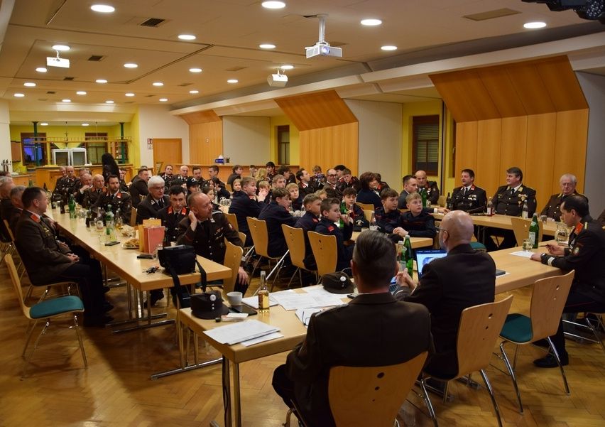Ein Saal mit Holzwänden und einer Bühne, in dem eine große Gruppe von Menschen in Uniformen an Tischen sitzt, möglicherweise in einer Besprechung. Flaschen, Papiere und Hüte sind auf den Tischen.