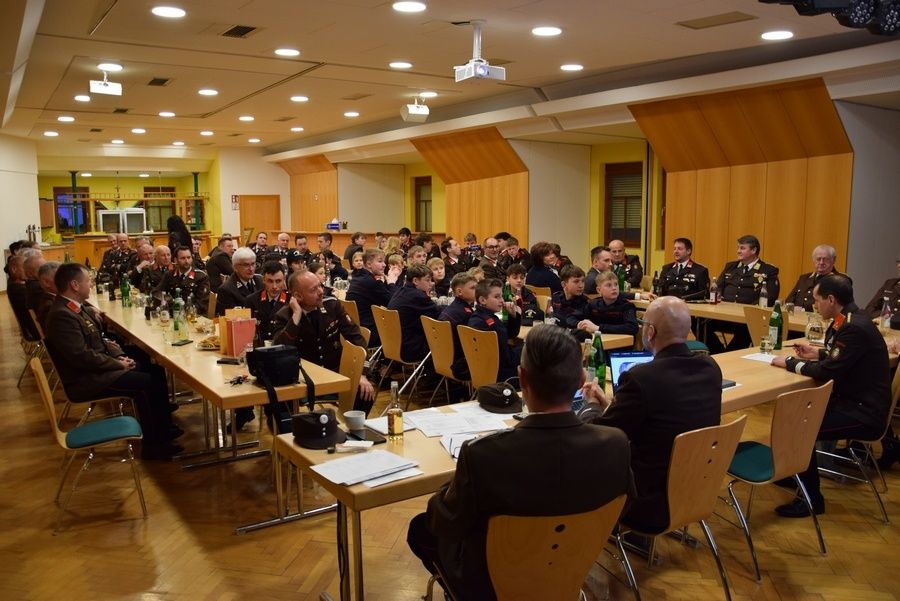 Ein Saal mit Holzwänden und einer Bühne, in dem eine große Gruppe von Menschen in Uniformen an Tischen sitzt, möglicherweise in einer Besprechung. Flaschen, Papiere und Hüte sind auf den Tischen.