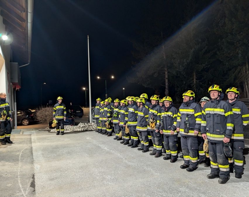 Eine Gruppe von Feuerwehrleuten steht in einer Reihe, bekleidet mit Helmen und Uniformen. Sie stehen auf einem Betonweg, und der Hintergrund zeigt einen dunklen Himmel mit Bäumen.