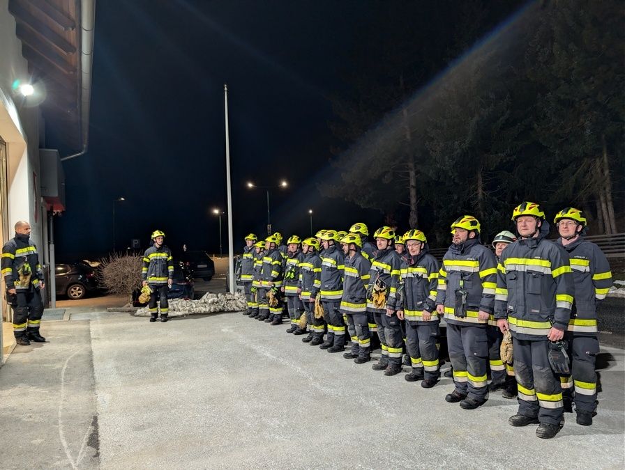 Eine Gruppe von Feuerwehrleuten steht in einer Reihe, bekleidet mit Helmen und Uniformen. Sie stehen auf einem Betonweg, und der Hintergrund zeigt einen dunklen Himmel mit Bäumen.