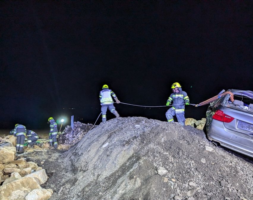 Mehrere Feuerwehrleute arbeiten nachts daran, ein Auto aus einem Haufen Steine zu ziehen. Sie tragen gelbe Helme und Sicherheitsanzüge.