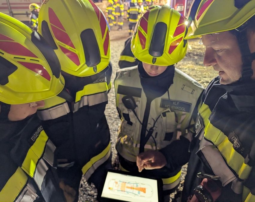 Eine Gruppe junger Feuerwehrleute in gelben Helmen und Uniformen versammelt sich um einen Mann, der auf einen Tablet-Bildschirm zeigt, wobei ein Feuerwehrwagen im Hintergrund zu sehen ist.