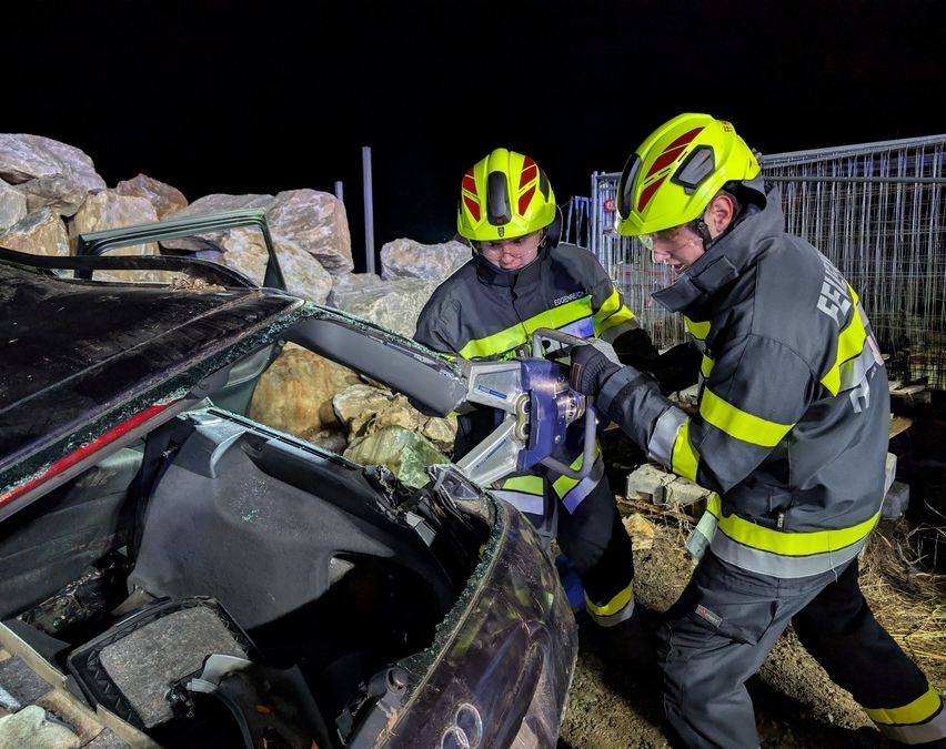 Zwei Feuerwehrleute verwenden Werkzeuge, um die beschädigte Tür eines verunglückten Autos aufzubrechen, mit Felsen und einem Metallzaun im Hintergrund.