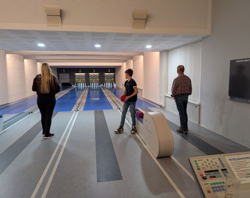 Drei Personen befinden sich in einer Bowlingbahn. Ein Mann bereitet sich darauf vor, einen roten Bowlingball zu werfen, während zwei andere zusehen.