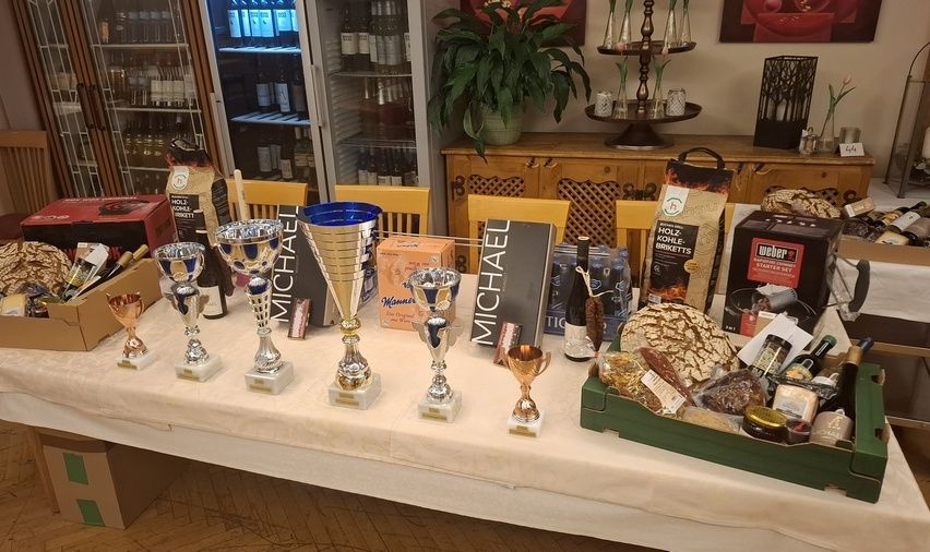 Ein Tisch ist mit Trophäen, Flaschen und verschiedenen Lebensmitteln beladen. Hinter dem Tisch befinden sich Regale mit Weinflaschen und einer Pflanze.