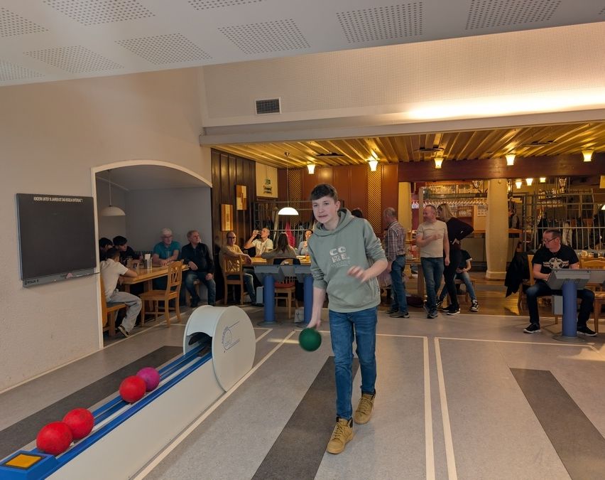 Ein junger Mann in einer Kapuzenjacke bereitet sich darauf vor, einen grünen Ball auf einer Bowlingbahn zu werfen. Hinter ihm sitzen Leute an Tischen und schauen zu.