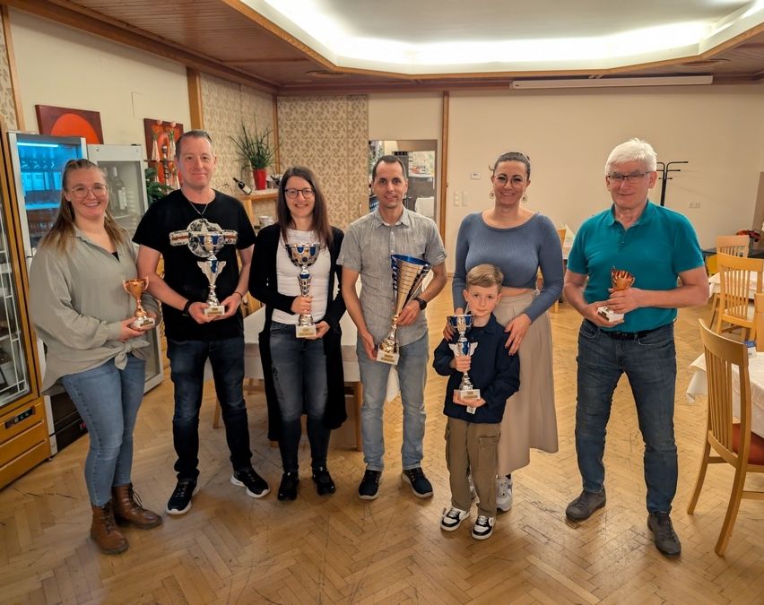 Eine Gruppe von Menschen steht in einem Raum, lächelt und hält Pokale. Ein Kind ist in der Mitte. Ein Kühlschrank, eine Pflanze und ein Bilderrahmen sind im Hintergrund.