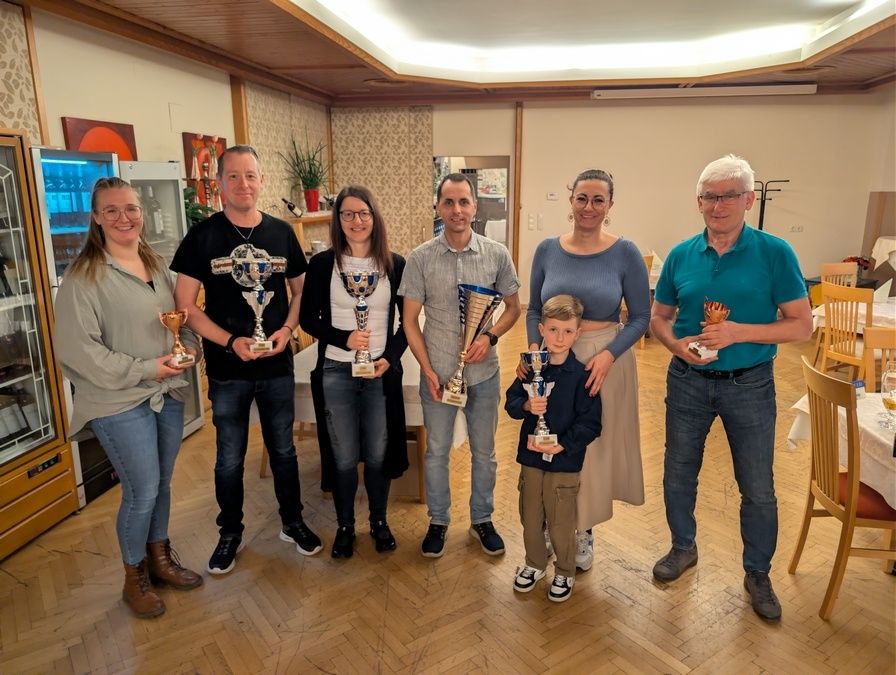 Eine Gruppe von Menschen steht in einem Raum, lächelt und hält Pokale. Ein Kind ist in der Mitte. Ein Kühlschrank, eine Pflanze und ein Bilderrahmen sind im Hintergrund.