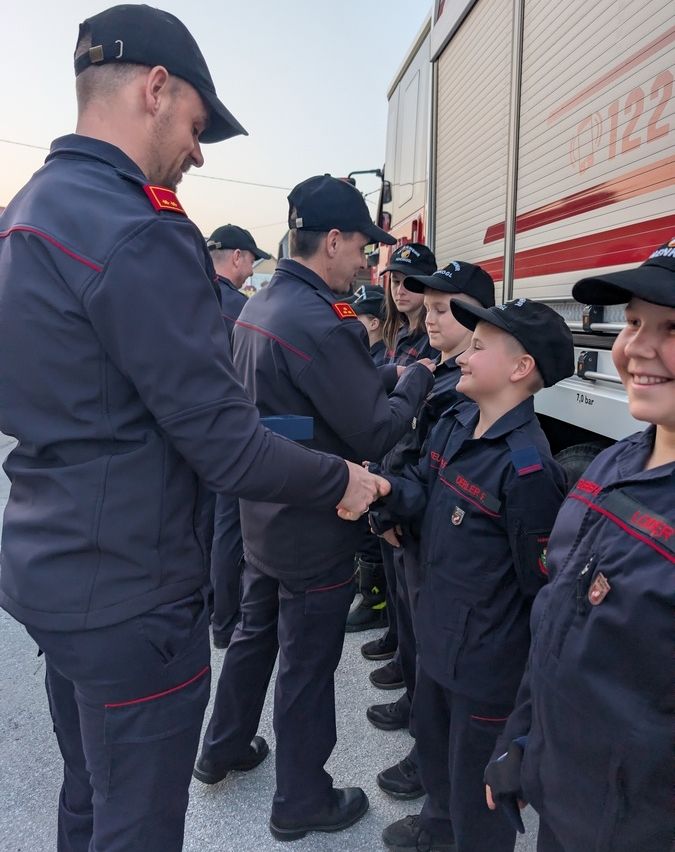 Eine Gruppe uniformierter Kinder gibt einem Feuerwehrmann draußen bei einem Feuerwehrauto die Hand.