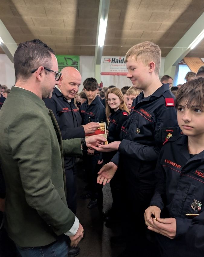 Eine Gruppe von Menschen in Uniformen steht in einem Raum. Ein Mann in einem grünen Mantel überreicht einem Jungen in einer schwarzen Uniform eine Medaille.