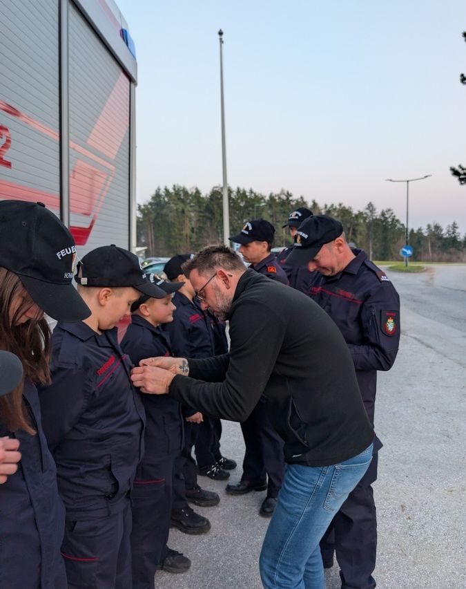 Ein Mann verteilt Auszeichnungen an mehrere junge Personen in Uniform, die vor einem Feuerwehrauto stehen.