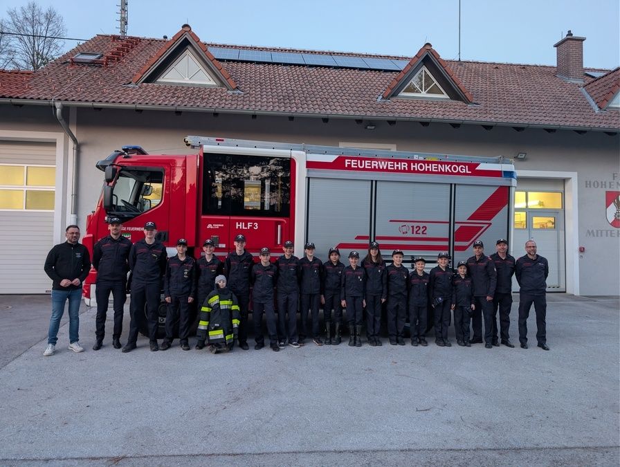 Eine Gruppe von Feuerwehrleuten und einem Kind steht vor einem Feuerwehrauto vor einem Gebäude mit Solarzellen auf dem Dach.