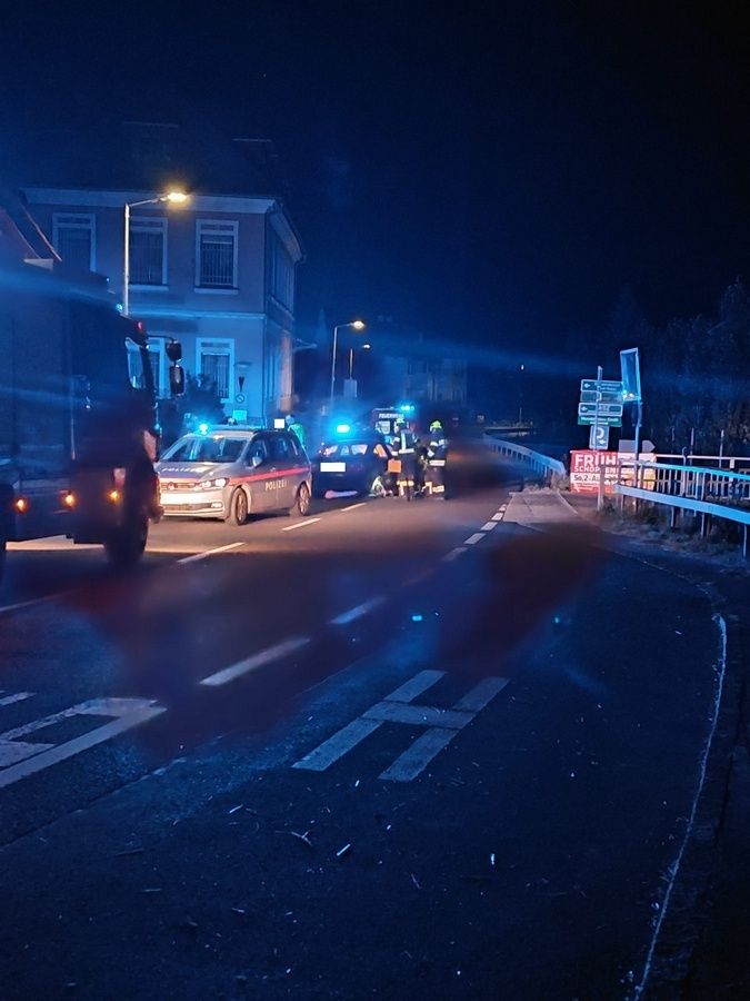 Notfallfahrzeuge, einschließlich eines Polizeiwagens, sind nachts auf einer Straße geparkt. Eine Gruppe von Menschen, möglicherweise Rettungskräfte, steht in der Nähe der Fahrzeuge. Der Bereich wird von den blinkenden Lichtern der Fahrzeuge beleuchtet.