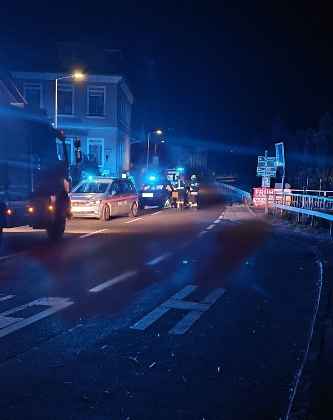 Notfallfahrzeuge, einschließlich eines Polizeiwagens, sind nachts auf einer Straße geparkt. Eine Gruppe von Menschen, möglicherweise Rettungskräfte, steht in der Nähe der Fahrzeuge. Der Bereich wird von den blinkenden Lichtern der Fahrzeuge beleuchtet.