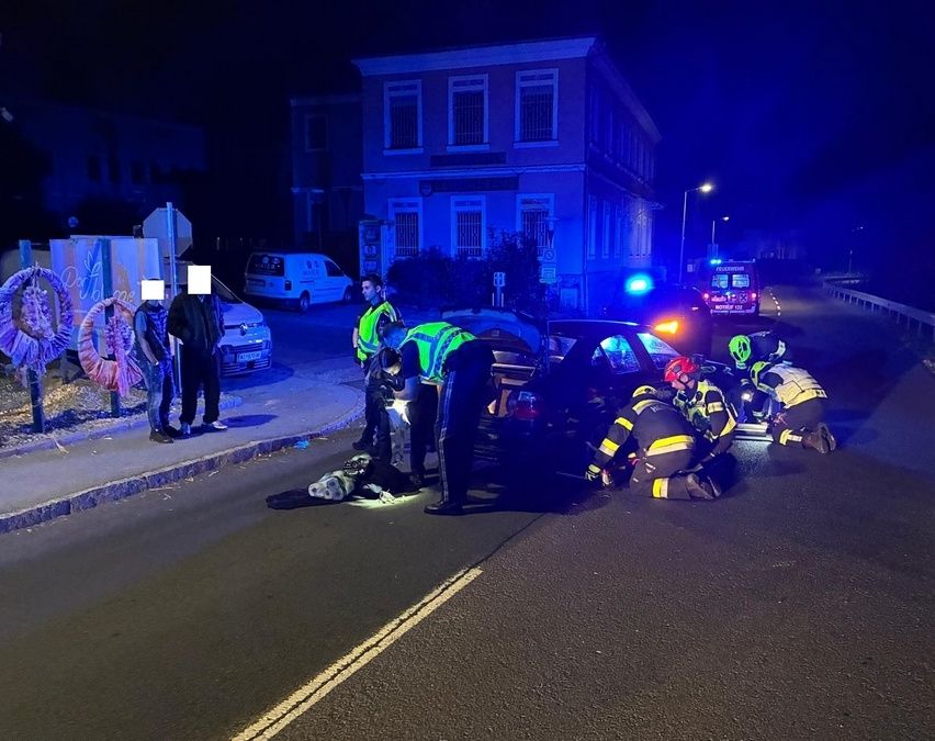 Rettungskräfte kümmern sich nachts auf einer Stadtstraße um einen Autounfall, mit Schaulustigen und Rettungsfahrzeugen sichtbar.