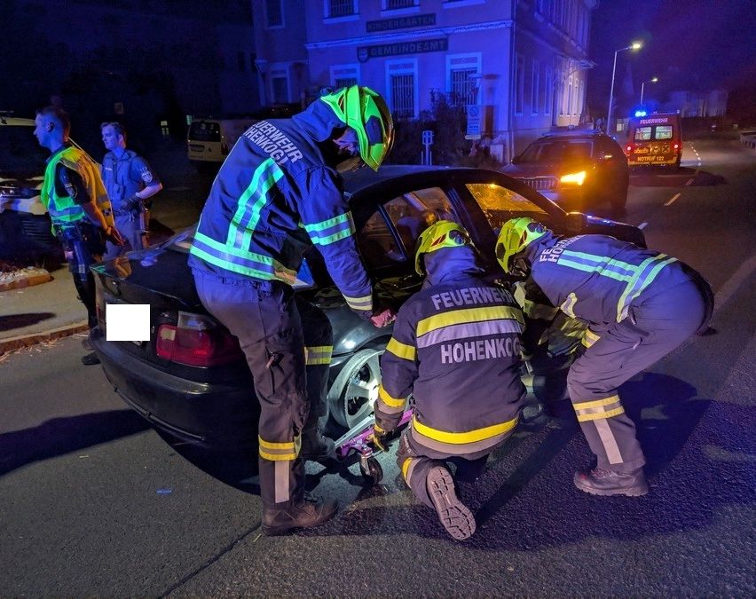 Feuerwehrleute in Schutzkleidung arbeiten an einem beschädigten Auto auf einer Straße bei Nacht. Ein Gebäude mit einem Schild ist im Hintergrund zu sehen.