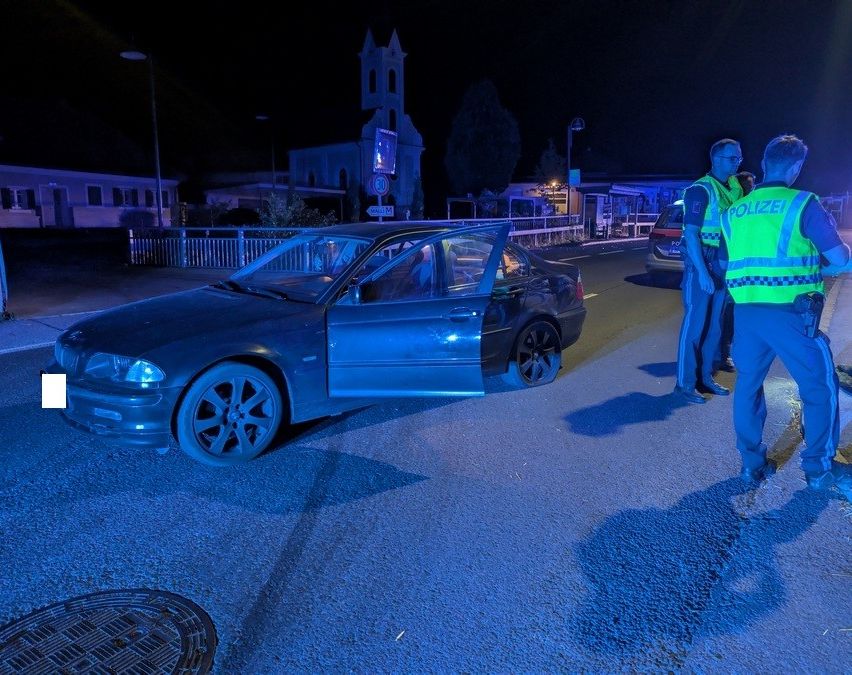 Zwei Polizeibeamte in reflektierenden Westen stehen neben einem beschädigten Auto auf einer Straße bei Nacht. Die Autotür ist offen, und der Fahrer ist im Inneren. Eine Straßenlaterne und ein Gebäude sind im Hintergrund zu sehen.
