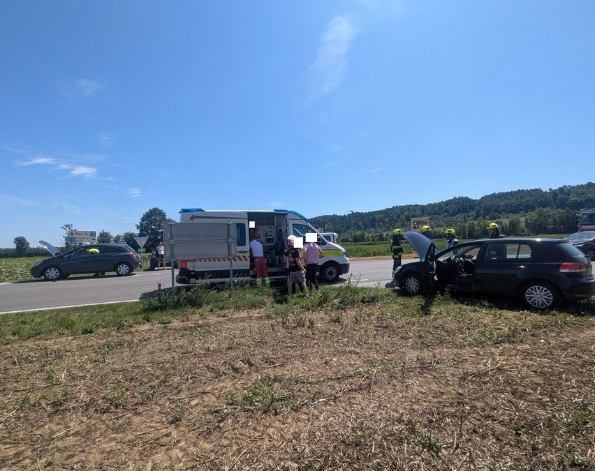 Ein Krankenwagen und ein Auto sind am Straßenrand geparkt, mit Leuten, die herumstehen. Der Kofferraum des Autos ist geöffnet.