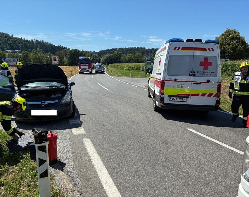 Ein Krankenwagen ist am Straßenrand geparkt, mit offenen Hecktüren. Ein Auto mit hochgeklappter Motorhaube steht in der Nähe. Eine Person sitzt mit einem Helm auf dem Boden.