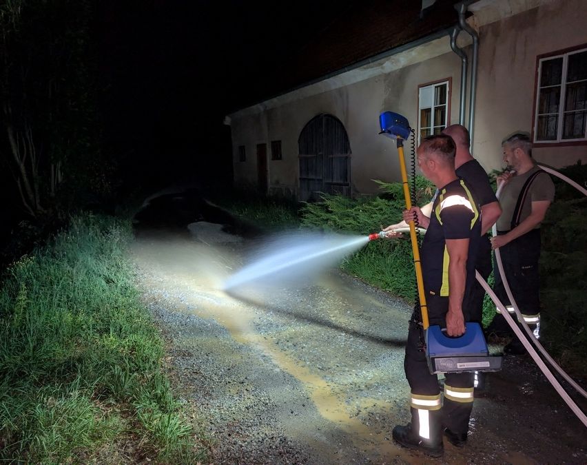 Drei Feuerwehrleute sprühen nachts Wasser auf eine Straße, mit einem Gebäude im Hintergrund.