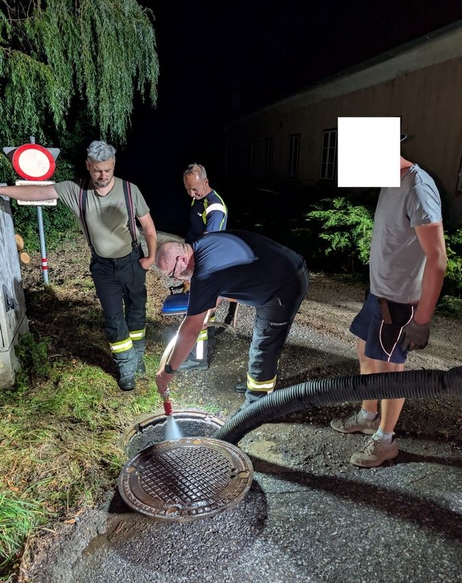 Vier Männer arbeiten nachts draußen, einer reinigt einen Schachtdeckel mit einem Schlauch.