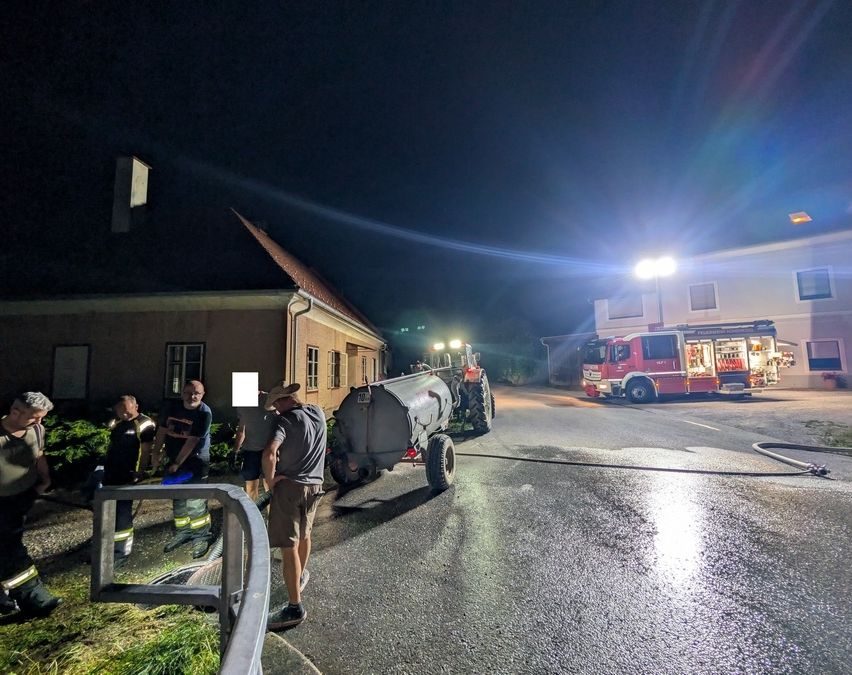 Ein Feuerwehrauto steht nachts am Straßenrand, mit Feuerwehrleuten, die um einen Traktor herumstehen. Der Traktor ist mit einem großen Tank verbunden, und die Szene wird von den Lichtern des Trucks beleuchtet.