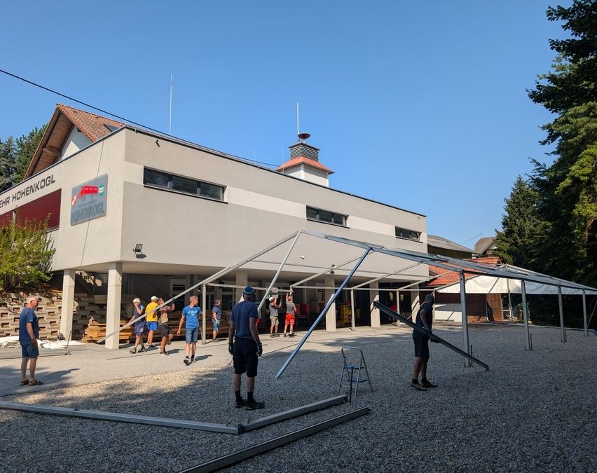 Eine Gruppe von Menschen baut ein Zelt vor einem Gebäude mit dem Schriftzug 'KOGL' auf. Das Zelt wird von zwei Personen zusammengebaut, während andere stehen und zusehen. Das Gebäude hat ein rotes Vordach und mehrere Fenster. Der Himmel ist klar und blau.
