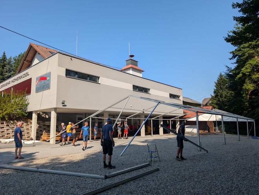 Eine Gruppe von Menschen baut ein Zelt vor einem Gebäude mit dem Schriftzug 'KOGL' auf. Das Zelt wird von zwei Personen zusammengebaut, während andere stehen und zusehen. Das Gebäude hat ein rotes Vordach und mehrere Fenster. Der Himmel ist klar und blau.
