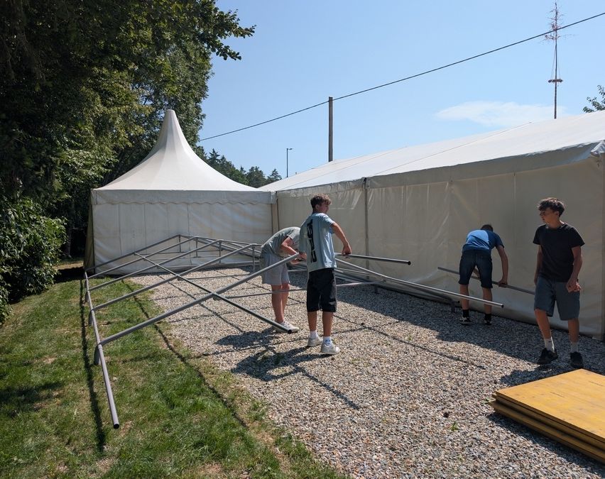 Drei Personen bauen ein großes weißes Zelt auf einer Kiesfläche auf. Eine Person beugt sich vor, während die anderen Metallstangen halten. Bäume und ein Strommast sind im Hintergrund zu sehen.