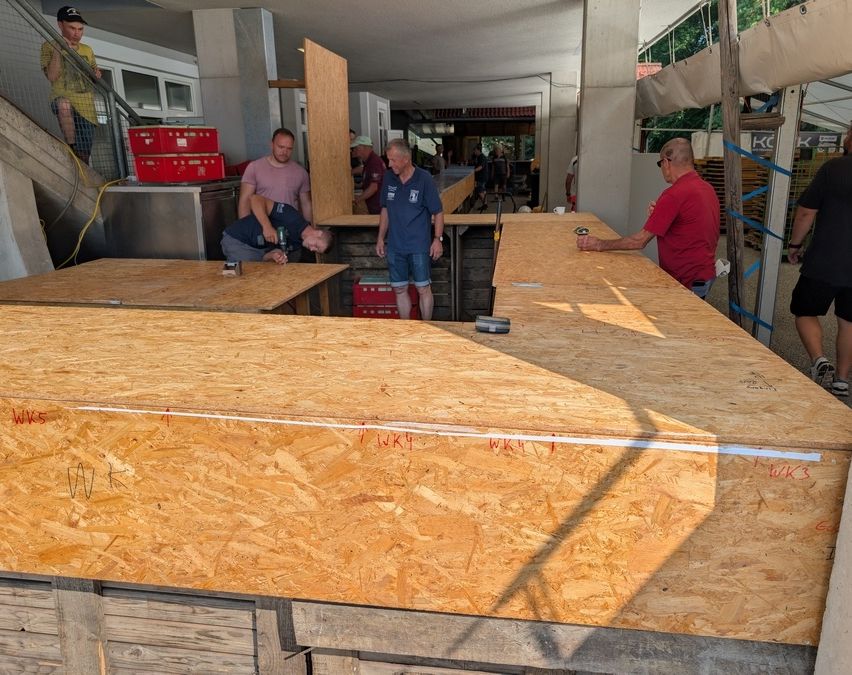 Eine Gruppe von Männern arbeitet an einer großen Holzstruktur, möglicherweise einer Bar, in einer Außenumgebung. Ein Mann beugt sich, ein anderer steht und ein dritter hält eine Dose. Im Hintergrund befinden sich Holzkisten und eine Treppe.