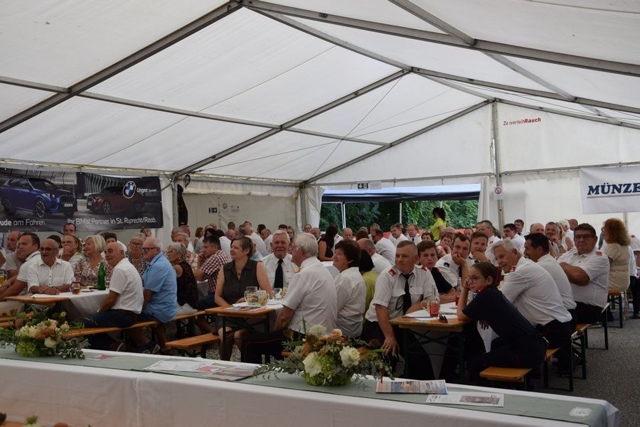 Eine Gruppe von Menschen in weißen Hemden und Krawatten sitzt an Tischen unter einem weißen Zelt mit Blumen und Getränken. Ein BMW-Banner ist links angebracht.