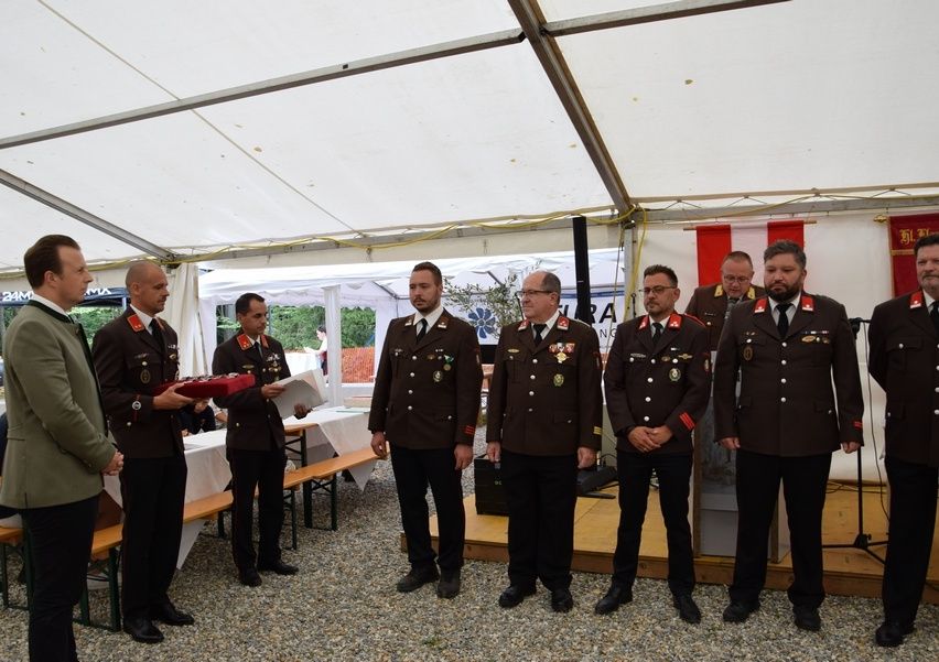 Eine Gruppe von Männern in Militäruniformen steht in einer Reihe unter einem Zelt, möglicherweise während einer Zeremonie. Sie tragen alle Medaillen und Krawatten. Ein Mann hält eine rote Mappe. Hinter ihnen steht ein Tisch mit einem weißen Tuch.