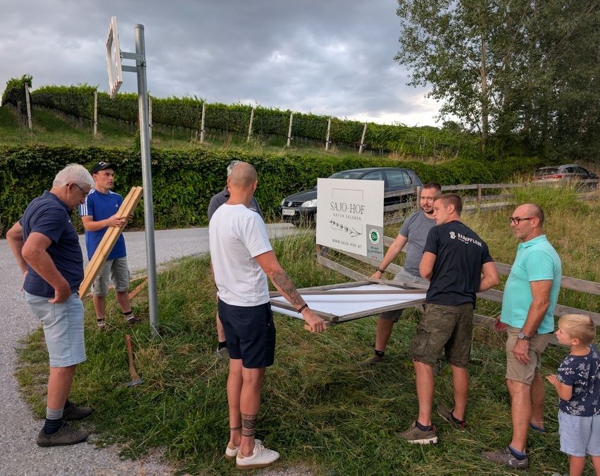 Eine Gruppe von Männern arbeitet an einem Schild in einem Grasbereich. Ein Mann hält eine Holzplanke und ein anderer eine Metallplatte. Ein Auto ist am Straßenrand geparkt.