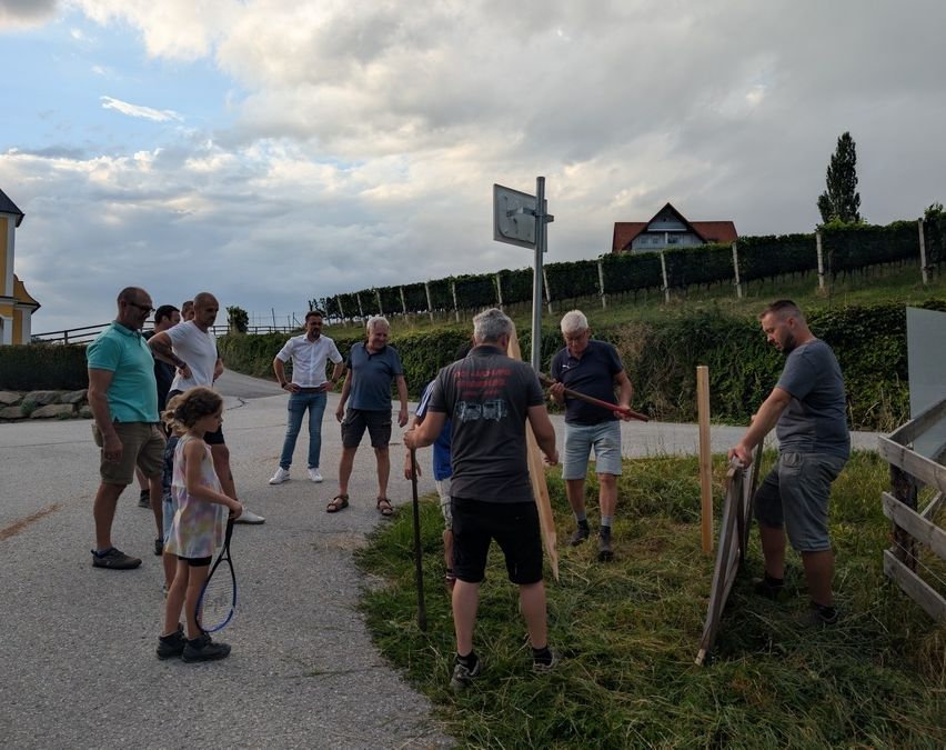 A group of people are standing on a road, some are holding sticks and appear to be working on the grass. A house and a basketball hoop are visible in the background.