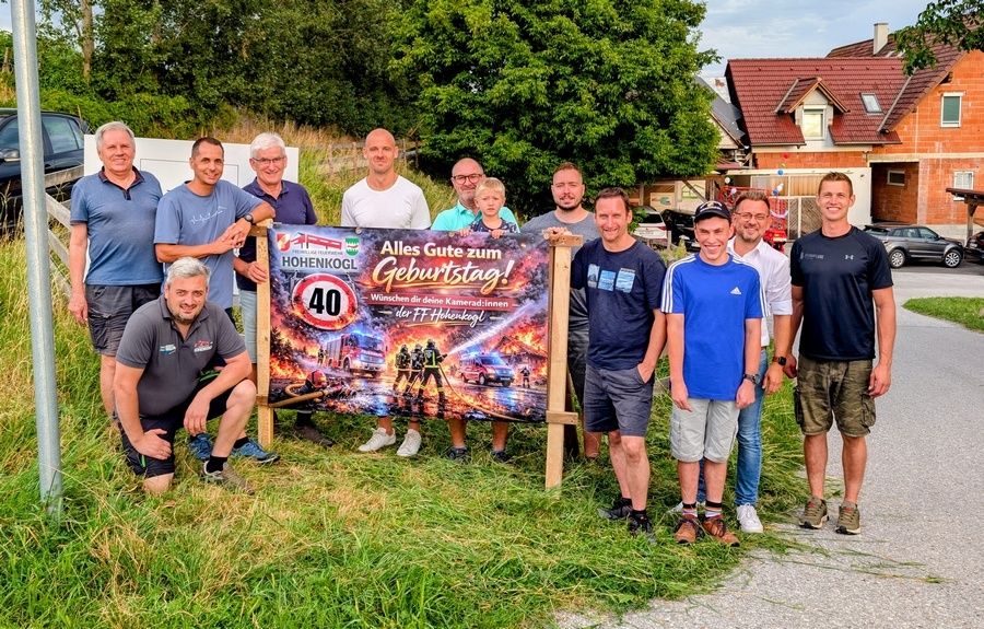 A group of men pose for a photo in front of a birthday banner with a fire truck image and the number 40. They are all smiling and standing in a row.