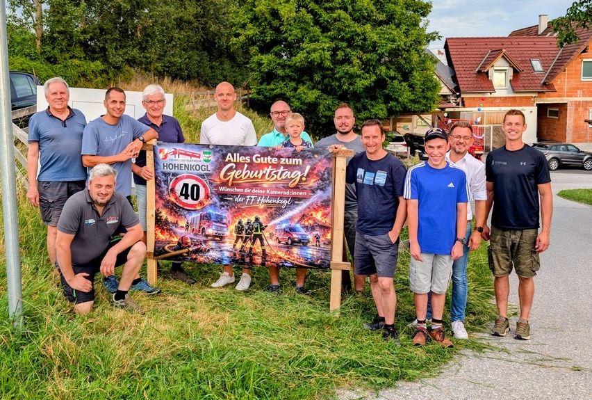 Eine Gruppe von Männern posiert für ein Foto vor einem Geburtstagsbanner mit einem Feuerwehrwagen-Bild und der Nummer 40. Sie alle lächeln und stehen in einer Reihe.