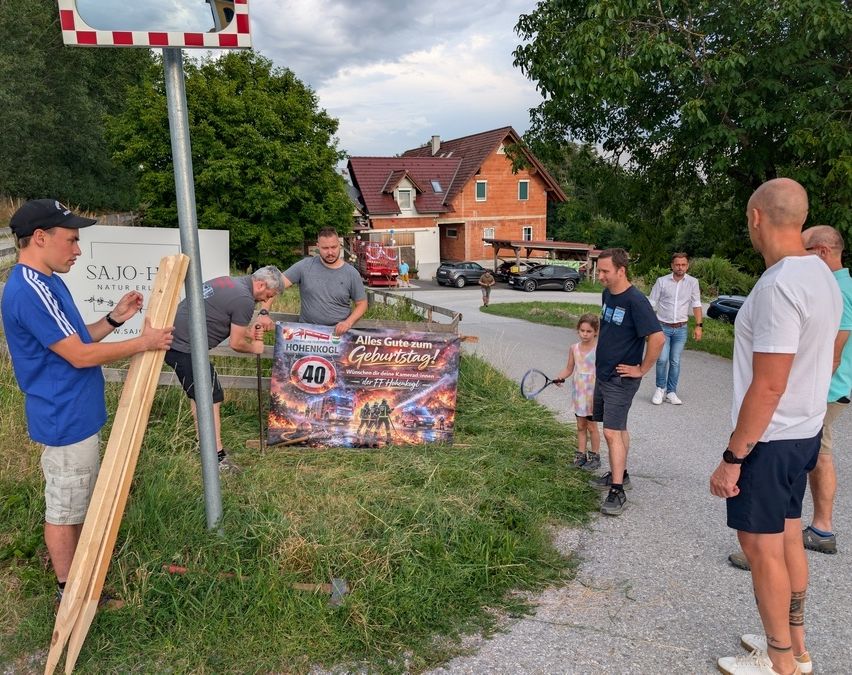 A group of people are gathered around a banner celebrating a 40th birthday. A man is holding a wooden plank, and another man is fixing something on a pole. A girl is playing with a tennis racket. A house and cars are in the background.