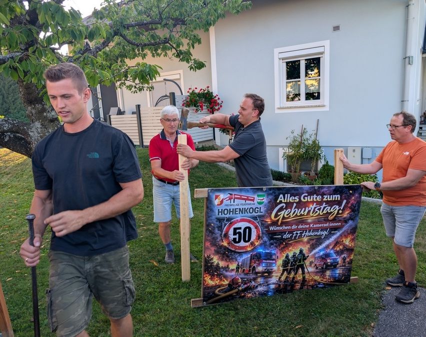 Four men are standing in a yard, one holding a sign that reads 'Alles Gute zum Geburtstag!' with a fire truck and firefighters on it. They are standing in front of a house with a tree and flowers.