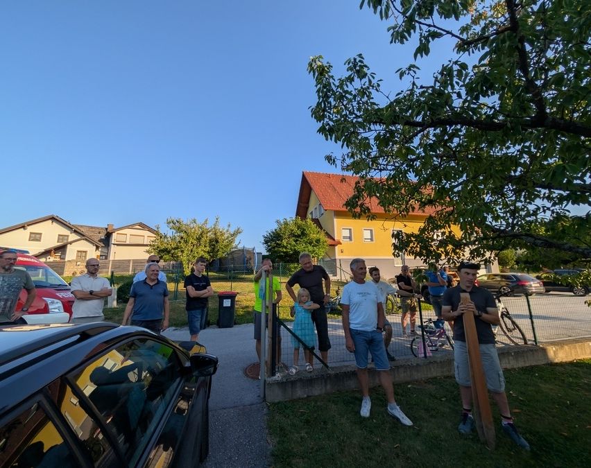 A group of people stands near a yellow house with a red roof, some are wearing hats and sneakers, while others are standing by a fence with bicycles. A car is parked on the side.