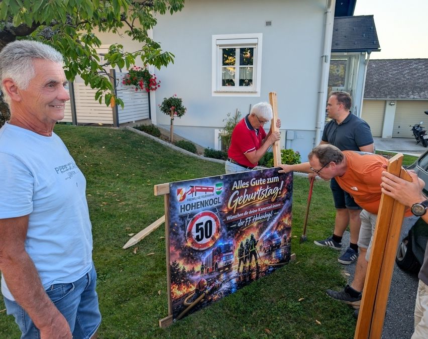 Vier Männer stehen in einem Garten, zwei von ihnen reparieren ein Schild, das 'Hohenkohl 50' steht. Der Mann auf der linken Seite lächelt und schaut zur Kamera. Der Mann in der Mitte trägt ein rotes Hemd und eine Brille. Der Mann auf der rechten Seite trägt ein orangefarbenes Hemd und eine Brille. Hinter ihnen steht ein Haus mit einem Fenster und einer Pflanze.
