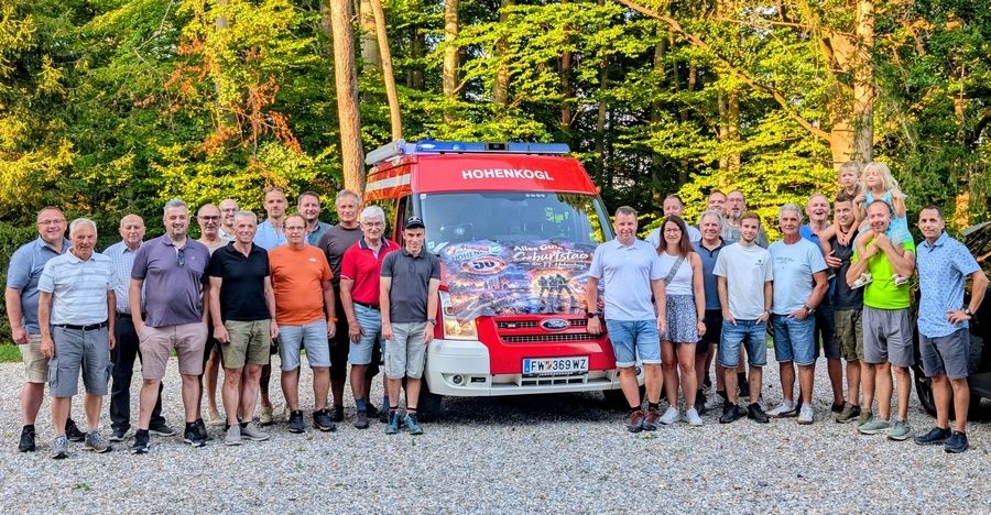 A group of people, including men and women, are posing for a photo in front of a red fire truck with the word Hohenkogl on it. They are all smiling and seem to be having a good time.