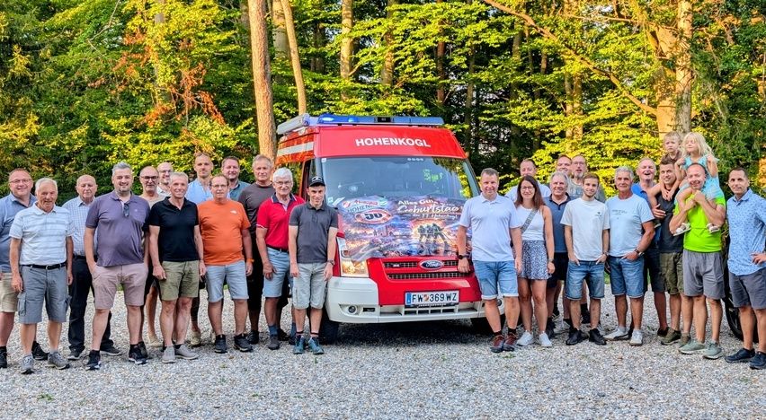 Eine Gruppe von Menschen, einschließlich Männer und Frauen, posiert für ein Foto vor einem roten Feuerwehrauto mit dem Wort Hohenkogl darauf. Sie alle lächeln und scheinen eine gute Zeit zu haben.